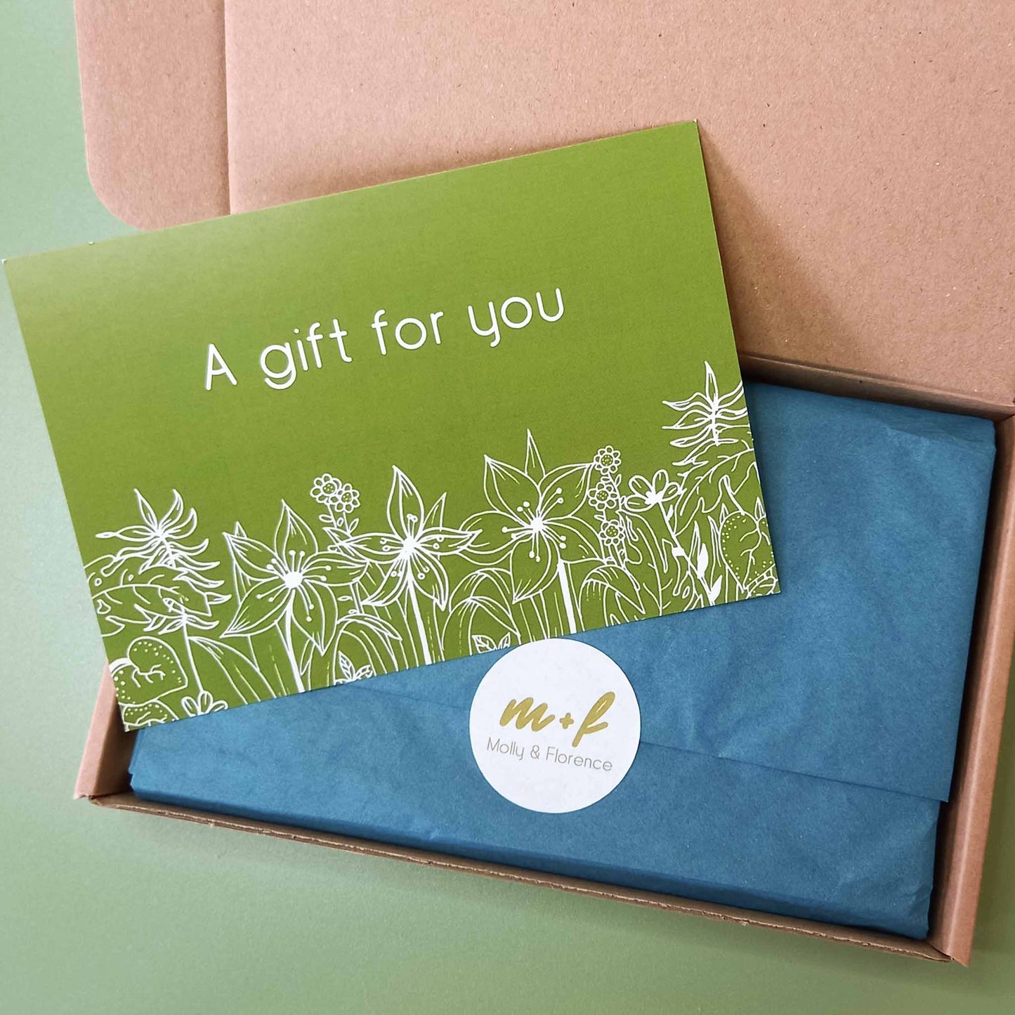 An open Molly and Florence gift box with blue tissue paper sealed with a branded sticker and a green gift note postcard