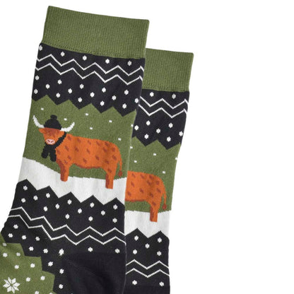 Close-up of a pair of green and black socks featuring snowflakes and a highland cow wearing a hat and scarf