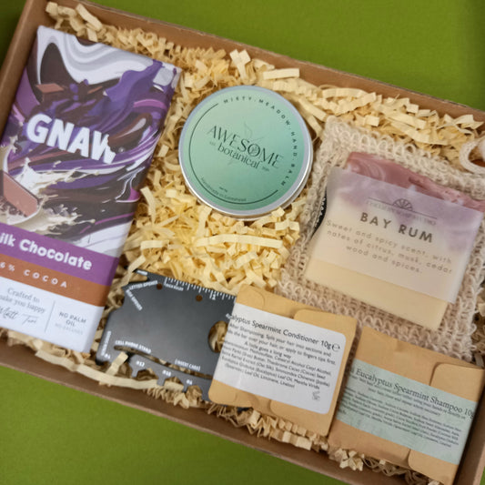 Gift box with various products including a bar of soap, a solid hand balm, and a chocolate on a green background