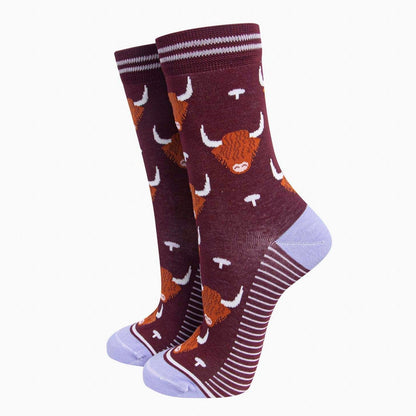 A pair of crew socks featuring a pattern of orange highland cow heads with white horns on a dark burgundy background, with lilac stripes on the cuffs and heels, and lilac toes and heels, shown against a plain white background.