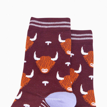 A pair of crew socks in dark burgundy with an orange and white highland cow pattern, featuring lilac stripes at the cuffs and a light lavender heel and toe cap.